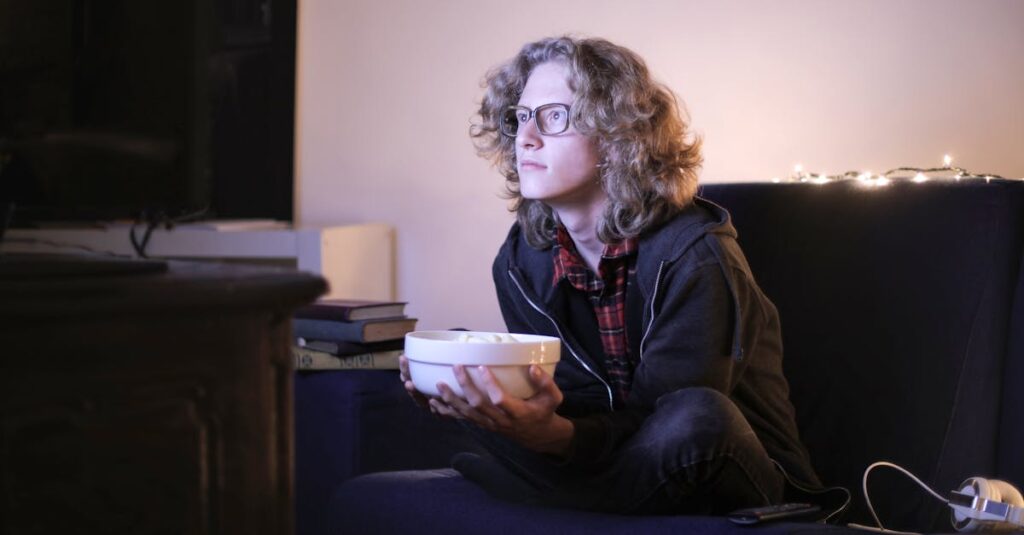 Young man with glasses watching television with focus while holding a snack bowl in a cozy living room setting.