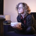Young man with glasses watching television with focus while holding a snack bowl in a cozy living room setting.